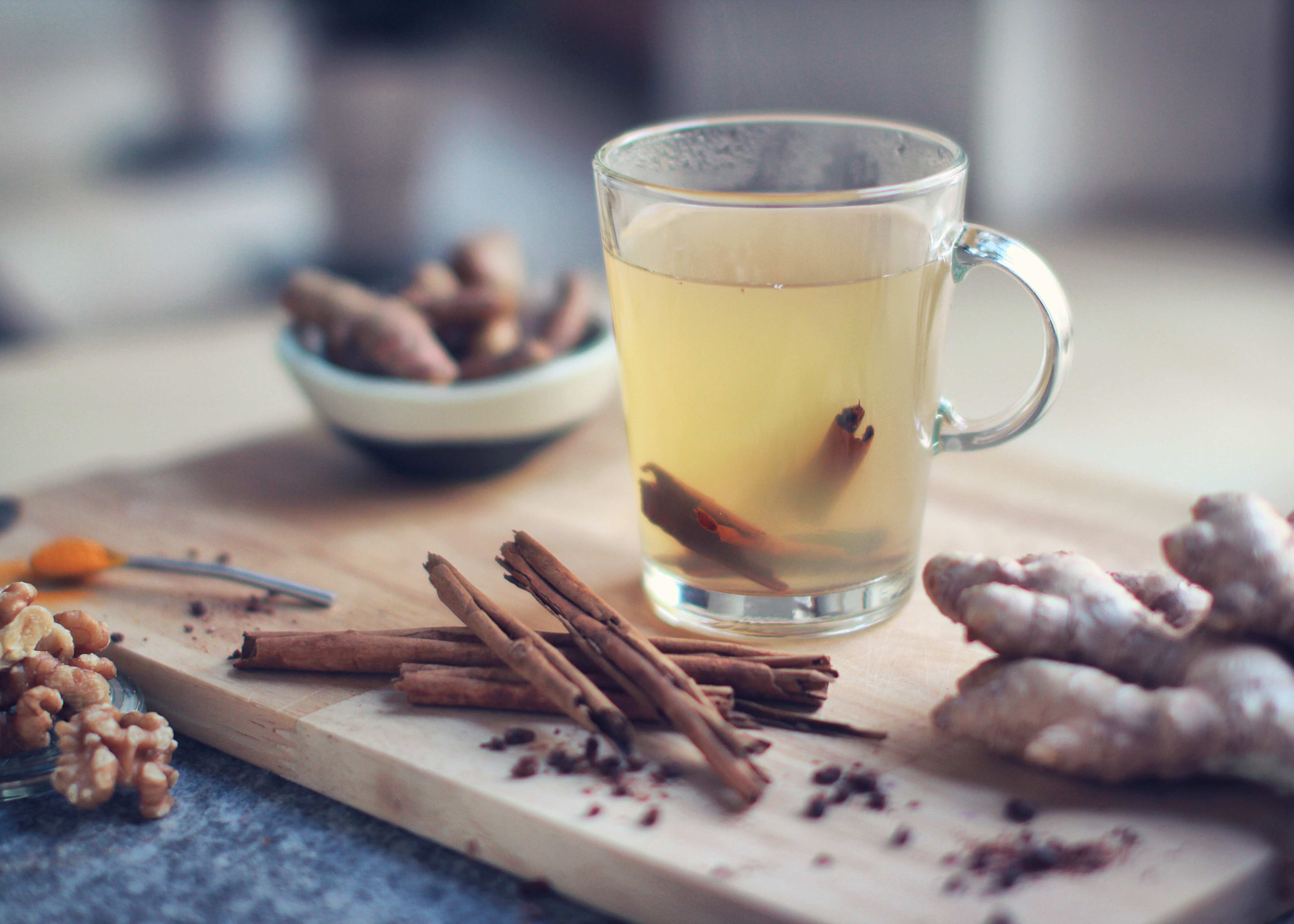 Zázvorový čaj ve sklenici s uchem na dřevěném kuchyňském prkénku. Kolem jsou položené kousky zázvoru a ořechy.